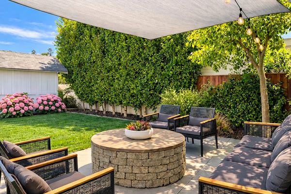 Backyard patio with stone fire pit, lounge seating, shade sail, string lights, and lush green hedging