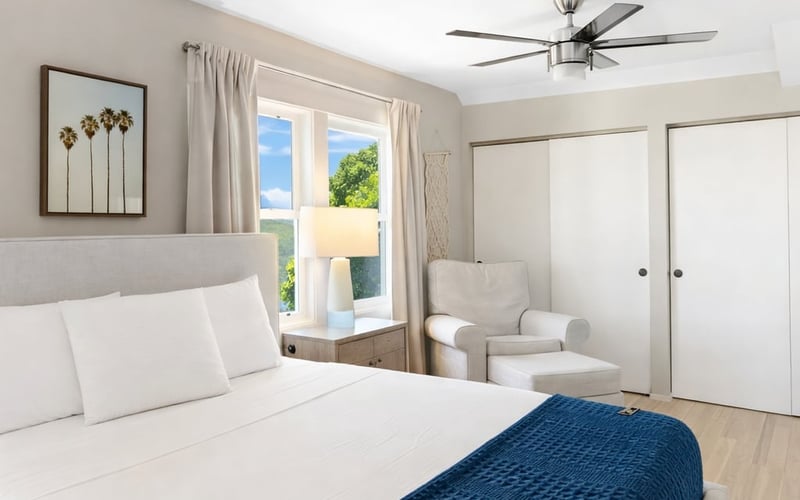 Bedroom with a navy throw across white bedding, a ceiling fan, reading chair and sunlit window