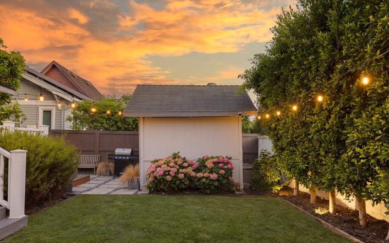 Backyard lawn at sunset, edged with string lights and a garden shed under an orange sky