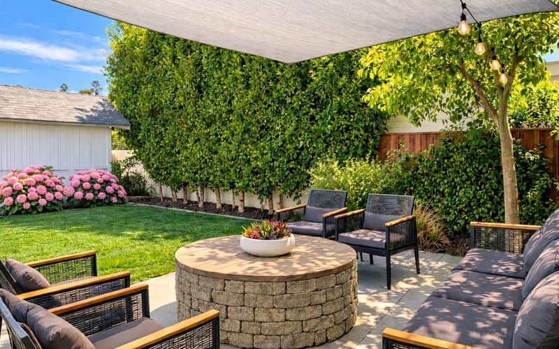 Backyard patio with stone fire pit, lounge seating, shade sail, ivy-covered walls, and pink flowering shrubs