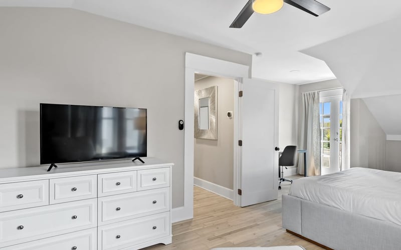 Upstairs bedroom with a wide TV on a white dresser, ceiling fan, ottoman and a desk nook beyond
