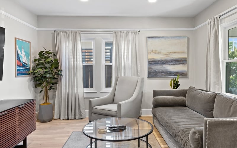 Living room with a grey sectional sofa, two armchairs, a round glass coffee table and coastal artwork