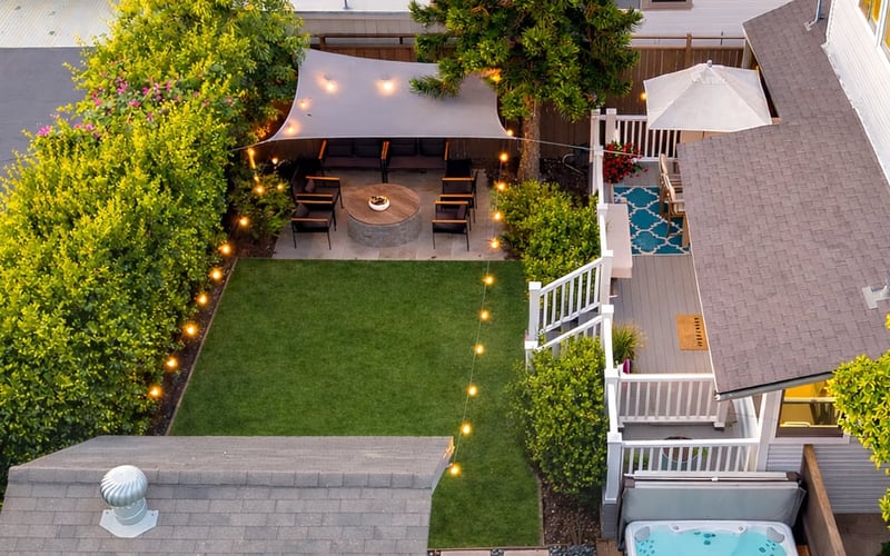 Aerial view of the fenced backyard at dusk with a bistro-lit dining pergola, lawn and hot tub